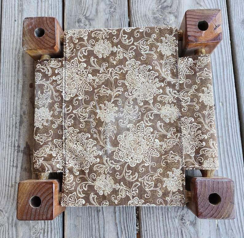 May include: A square footstool featuring a brown fabric top with a cream-colored floral damask pattern. The stool is supported by four thick, dark brown wooden legs, each with a circular hole drilled through the center of the top surface.