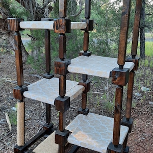 May include: A multi-level cat tree constructed from dark stained wood posts and square blocks. It features four platforms covered in a light beige fabric with a white geometric pattern, a vertical sisal rope scratching post, and a base with sisal matting.