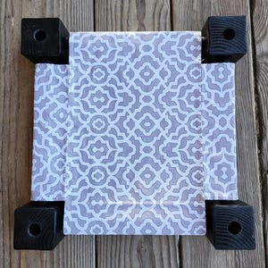 May include: A small square pet bed featuring a fabric center with a light gray and white geometric pattern. The bed is supported by four black wooden corner posts, each with a circular hole drilled into the top surface of the square block.