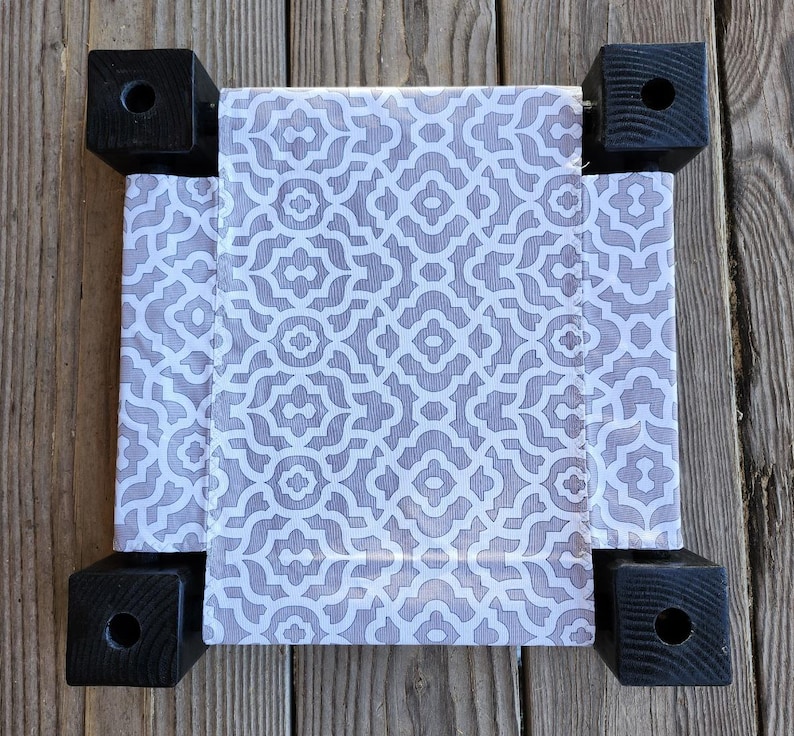 May include: A small square pet bed featuring a fabric center with a light gray and white geometric pattern. The bed is supported by four black wooden corner posts, each with a circular hole drilled into the top surface of the square block.