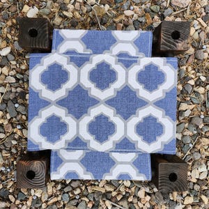 May include: A small stool featuring a blue and white fabric seat with a repeating quatrefoil pattern. The seat is supported by four dark brown, square-cut wooden legs, each with a circular hole drilled into the top surface of the post.