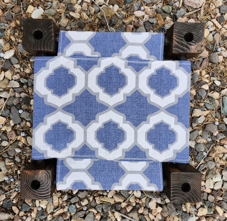 May include: A small stool featuring a blue and white fabric seat with a repeating quatrefoil pattern. The seat is supported by four dark brown, square-cut wooden legs, each with a circular hole drilled into the top surface of the post.