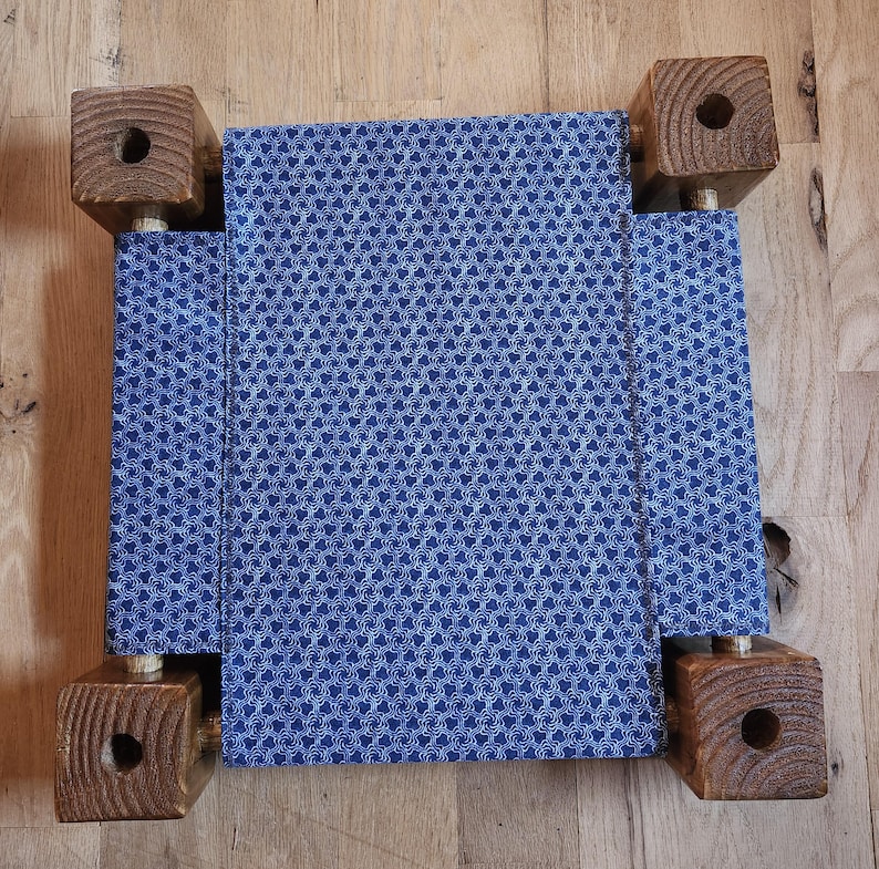 May include: A small pet bed featuring a rectangular blue fabric panel with a repeating white geometric pattern. The fabric is suspended between four square, dark brown wooden corner posts, each with a circular hole drilled into the top surface.