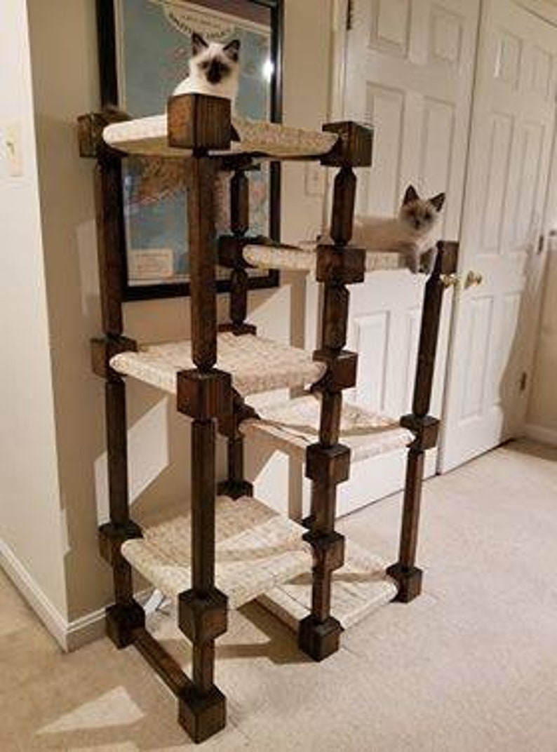 May include: A multi-level cat tower constructed from dark stained wooden posts and square connectors, featuring four beige fabric-covered platforms. Two light-colored Siamese cats are positioned on the top and second-highest levels.