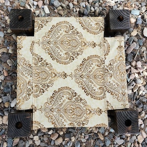 May include: A small, square, low-profile platform featuring four dark brown, stained wooden block legs with circular cutouts at the top corners. The center surface is covered in a cream-colored fabric with a repeating gold paisley pattern.