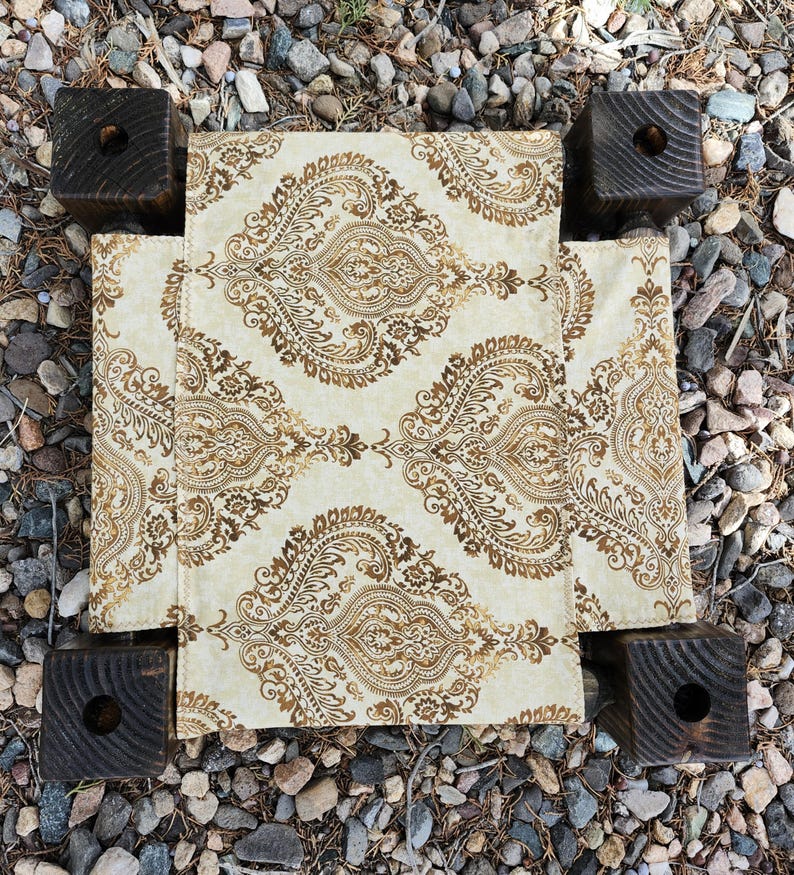 May include: A small, square, low-profile platform featuring four dark brown, stained wooden block legs with circular cutouts at the top corners. The center surface is covered in a cream-colored fabric with a repeating gold paisley pattern.