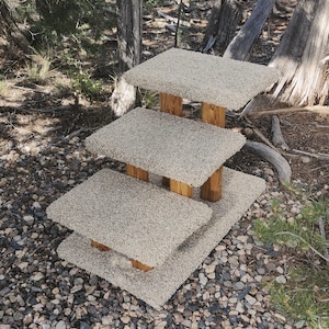 May include: A three-tiered pet staircase featuring rectangular platforms covered in beige, textured carpet. The structure is supported by vertical wooden posts with a natural grain finish, providing a stable base for each ascending step level.