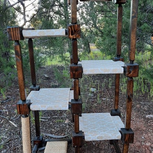 May include: A multi-level cat tree constructed from dark stained wood posts and square connectors. It features four elevated platforms covered in a light beige fabric with a white geometric pattern, plus a sisal scratching post and base pad.