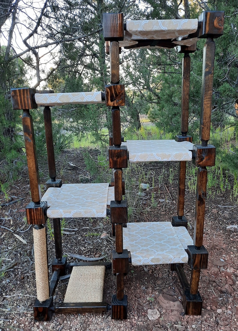 May include: A multi-level cat tree constructed from dark stained wood posts and square connectors. It features four elevated platforms covered in a light beige fabric with a white geometric pattern, plus a sisal scratching post and base pad.