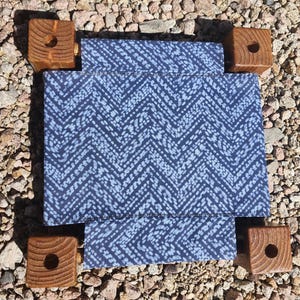 May include: A square fabric platform featuring a dark blue and white chevron pattern, supported by four thick, natural-toned wooden corner blocks. Each wooden block has a circular hole drilled into the center of its top surface.