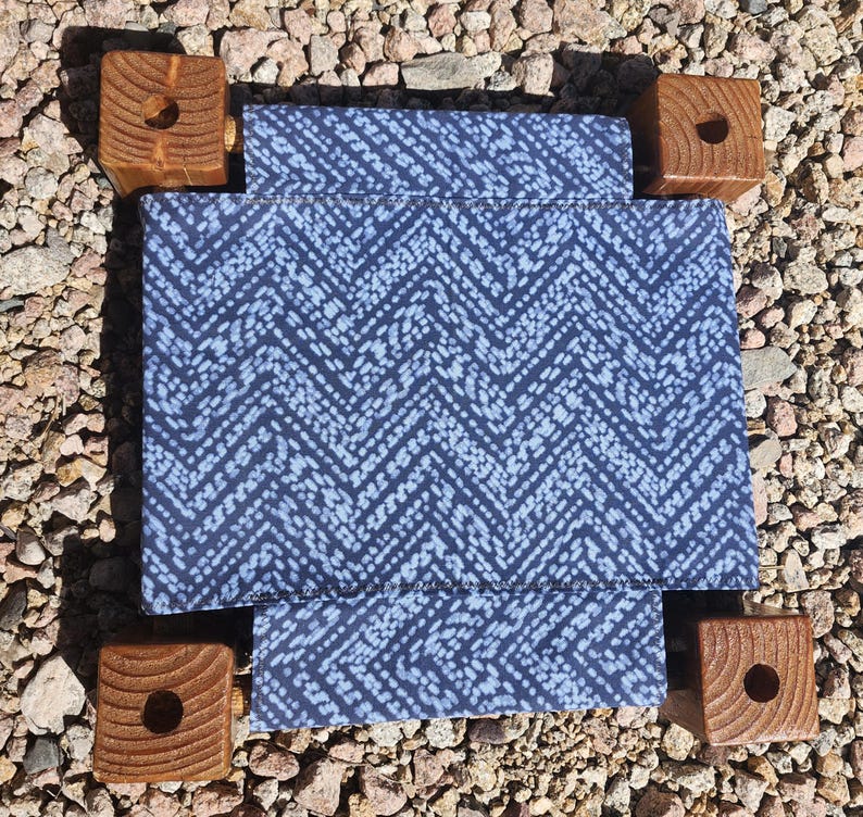 May include: A square fabric platform featuring a dark blue and white chevron pattern, supported by four thick, natural-toned wooden corner blocks. Each wooden block has a circular hole drilled into the center of its top surface.