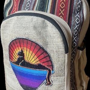 Könnte beinhalten: Ein beigefarbener Rucksack aus Hanf und Baumwolle mit einem schwarzen Reißverschluss und einem bunten gestickten Aufnäher mit einer Katze im Sonnenuntergang. Der Rucksack hat ein gestreiftes Muster auf der oberen Klappe. Der Aufnäher lautet "AKASHIC CONNECTIONS HEMP/COTTON MADE IN NEPAL".