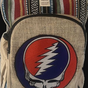 May include: A hemp and cotton backpack with a colorful woven top section and a large embroidered Steal Your Face logo. The main body is a natural beige color, with black zippers and straps. Made in Nepal.