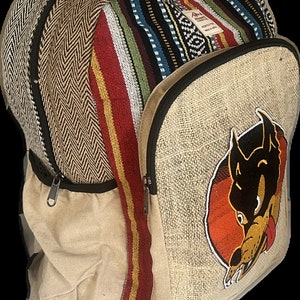 May include: A beige hemp backpack with a colourful striped pattern on the front flap. The backpack has a large zipped pocket on the front and a smaller zipped pocket on the side. The backpack also features an embroidered cartoon dog with a red tongue on the front.