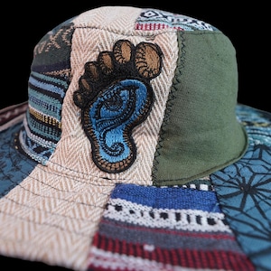 May include: A patchwork bucket hat featuring a variety of blue, green, and cream fabric panels with diverse woven patterns. The front panel displays a prominent embroidered patch of a blue foot with an eye design inside, outlined in dark brown thread.