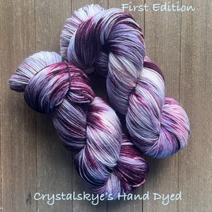 May include: Hand-dyed yarn skeins in shades of purple, white, and burgundy. The yarn is arranged in a loose, flowing pattern, with the text "First Edition" and "Crystalskye's Hand Dyed" visible. The yarn is on a wooden surface.