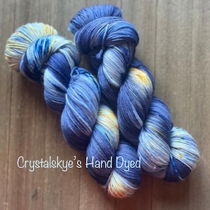 May include: Two skeins of hand-dyed yarn in shades of blue, white, and yellow. The yarn is twisted together and has a speckled appearance. The text "Crystalskye's Hand Dyed" is visible in the image.