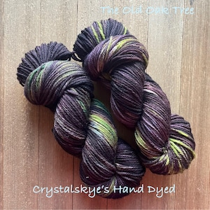 May include: Two skeins of hand-dyed yarn in shades of deep purple, green, and yellow. The yarn is arranged on a wooden surface. The text "The Old Oak Tree" and "Crystalskye's Hand Dyed" are visible.