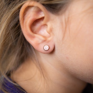 May include: A close-up of a person's ear with a small, round stud earring. The earring features a smiling face design in a light pink color. The background is blurred, with strands of blonde hair visible.