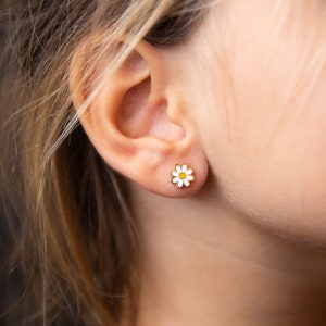 May include: A close-up of a person's ear with a small, gold daisy stud earring. The earring features a white enamel petal design with a yellow center. The earring is set against the skin, with blonde hair in the background.