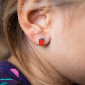 May include: A close-up of a child's ear with a small strawberry-shaped stud earring. The earring has a red body, green stem, and gold-colored outline. The earring is approximately 0.5 inches in height.