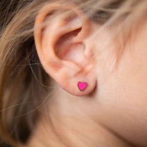 May include: A close-up of a person's ear with a pink heart-shaped stud earring. The earring has a gold-colored border. The ear is set against a blurred background.