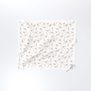 May include: White fabric with a repeating pattern of pink flowers, tan bunnies, and green leaves. The design is delicate and feminine, suitable for spring or Easter-themed projects. The fabric is folded and has a pair of scissors on the corner.