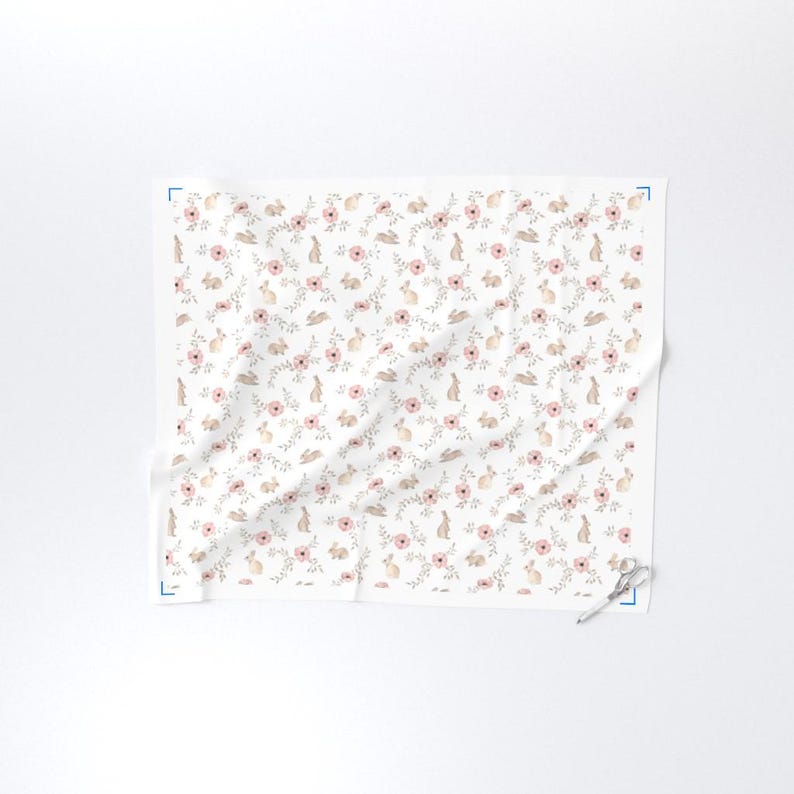May include: White fabric with a repeating pattern of pink flowers, tan bunnies, and green leaves. The design is delicate and feminine, suitable for spring or Easter-themed projects. The fabric is folded and has a pair of scissors on the corner.