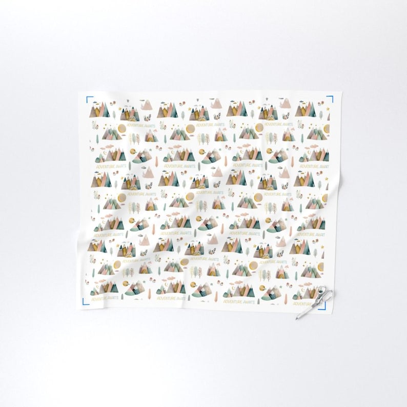 May include: White fabric with a repeating pattern of watercolor mountains, suns, clouds, and trees in muted colors. The text "ADVENTURE AWAITS" is printed throughout the design. The fabric is folded and has a pair of scissors in the corner.