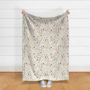 May include: A white blanket with a delicate floral pattern. The design features a dense arrangement of small flowers in shades of pink, purple, and peach, interspersed with green foliage. The blanket is held up, showcasing the full pattern.
