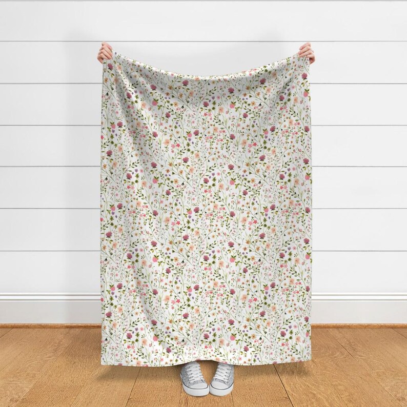 May include: A white blanket with a delicate floral pattern. The design features a dense arrangement of small flowers in shades of pink, purple, and peach, interspersed with green foliage. The blanket is held up, showcasing the full pattern.