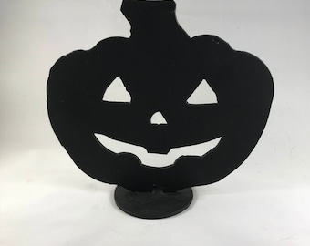 Op de afbeelding: Zwarte metalen Halloween-pompoensilhouet met een gesneden jack-o'-lantern-gezicht. De pompoen staat op een kleine ronde basis.