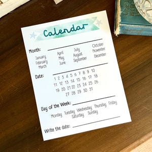 May include: A white calendar with the word "Calendar" in teal script. The calendar includes sections for the month, date, day of the week, and a space to write the date. The calendar is on a dark wooden surface.