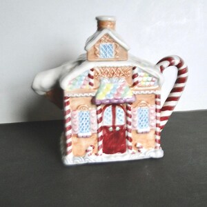 May include: A ceramic teapot in the shape of a gingerbread house. It has a red door, blue windows, and a candy cane handle. The roof is covered in white frosting and colourful candy decorations. A chimney is on top.