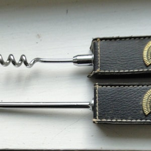 May include: A set of two black leather-covered bottle openers with gold-colored Roman helmet designs. The set includes a corkscrew and a spoon-shaped bottle opener.