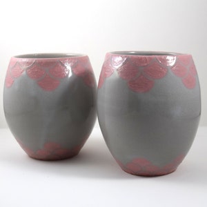 Puede incluir: Dos tazas de cerámica grises con un diseño festoneado rosa alrededor del borde superior.