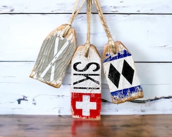 May include: Three wooden ski-themed ornaments hanging from a natural rope. One is gray with white ski graphics, the second has the word "SKI" and a red and white cross, and the third has black and blue diamond shapes.
