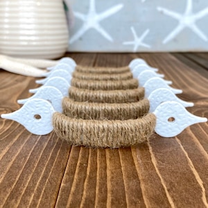 May include: A set of eight white ceramic drawer pulls with a rope-like design. The pulls are wrapped in natural jute twine and have a decorative, pointed shape.