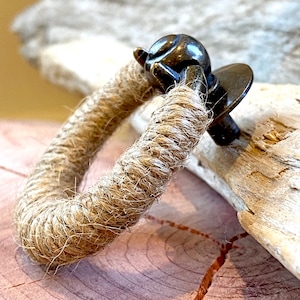 May include: A rustic bracelet featuring a thick, natural rope band. The rope is wrapped around a dark metal clasp with an ornate design. The bracelet rests on a wooden surface, showcasing its texture and earthy tones.