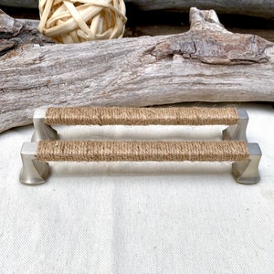 May include: Two silver cabinet pulls with a natural jute rope wrapped around the center. The pulls are on a white fabric background.