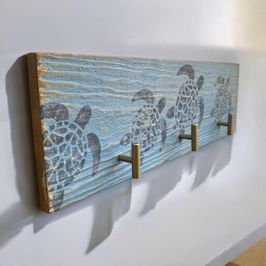May include: A wooden wall-mounted coat rack with a blue and white painted design of sea turtles. The rack has three gold metal hooks.