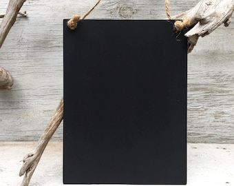 May include: A black chalkboard sign with a rustic twine hanger. The sign is hanging from a piece of driftwood against a white wood background.