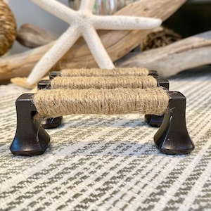 May include: Three brown metal drawer pulls wrapped in natural twine. The pulls are on a white and gray patterned surface.