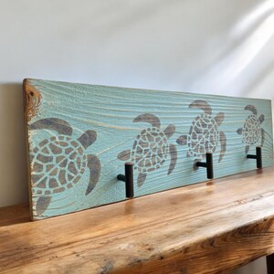 May include: A wooden wall-mounted coat rack with three hooks. The rack is painted a light blue color and features a stencil of three sea turtles in a row. The hooks are black metal.