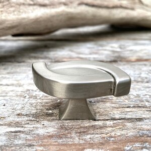 May include: A brushed nickel cabinet knob featuring a smooth, elongated, S-shaped top surface with rounded edges and a square-tapered base. The metal hardware has a matte, metallic finish and a curved, horizontal profile that extends outward.