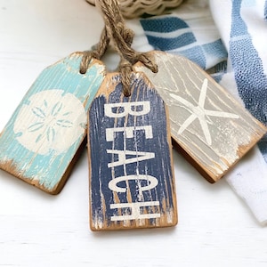 May include: Three wooden tags with a nautical theme. The tags are painted in blue, gray, and white with a starfish and seashell design. The middle tag says "BEACH" in white letters.