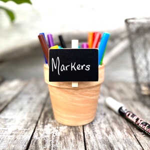 May include: A terracotta pot filled with colorful markers, labeled with a small black chalkboard sign that reads "Markers".