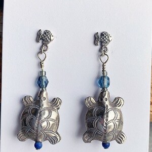 Op de afbeelding: Zilveren schildpad oorbellen met blauwe glazen kralen en een zilveren vis charme.  BEADS BY BURCH is gedrukt op de witte kaart.