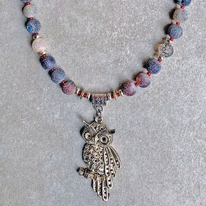 Puede incluir: Un collar hecho de cuentas de ágata en tonos grises, rojos y blancos con un colgante de búho plateado.