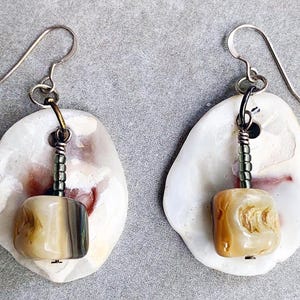 May include: A pair of earrings with a white shell and a brown and white bead. The earrings are made with silver wire and have a silver hook.